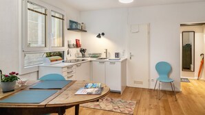 Una cocina compacta con armarios blancos, una mesa de comedor con sillas y un ventanal con persianas.