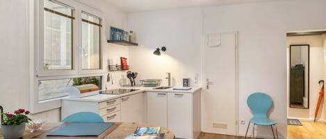 A compact kitchen with white cabinets, a dining table with chairs, and a small window with blinds.