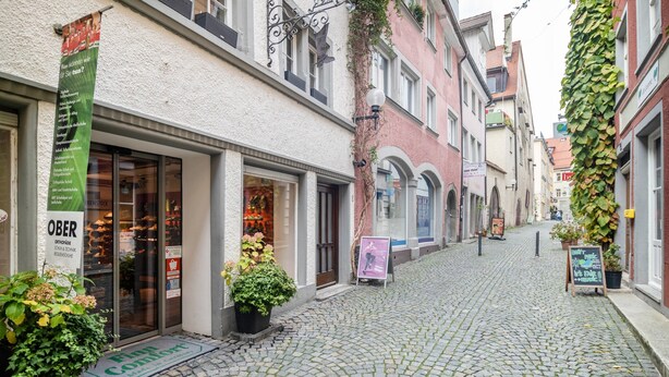 Una calle adoquinada con tiendas, un estandarte verde y plantas en macetas.
