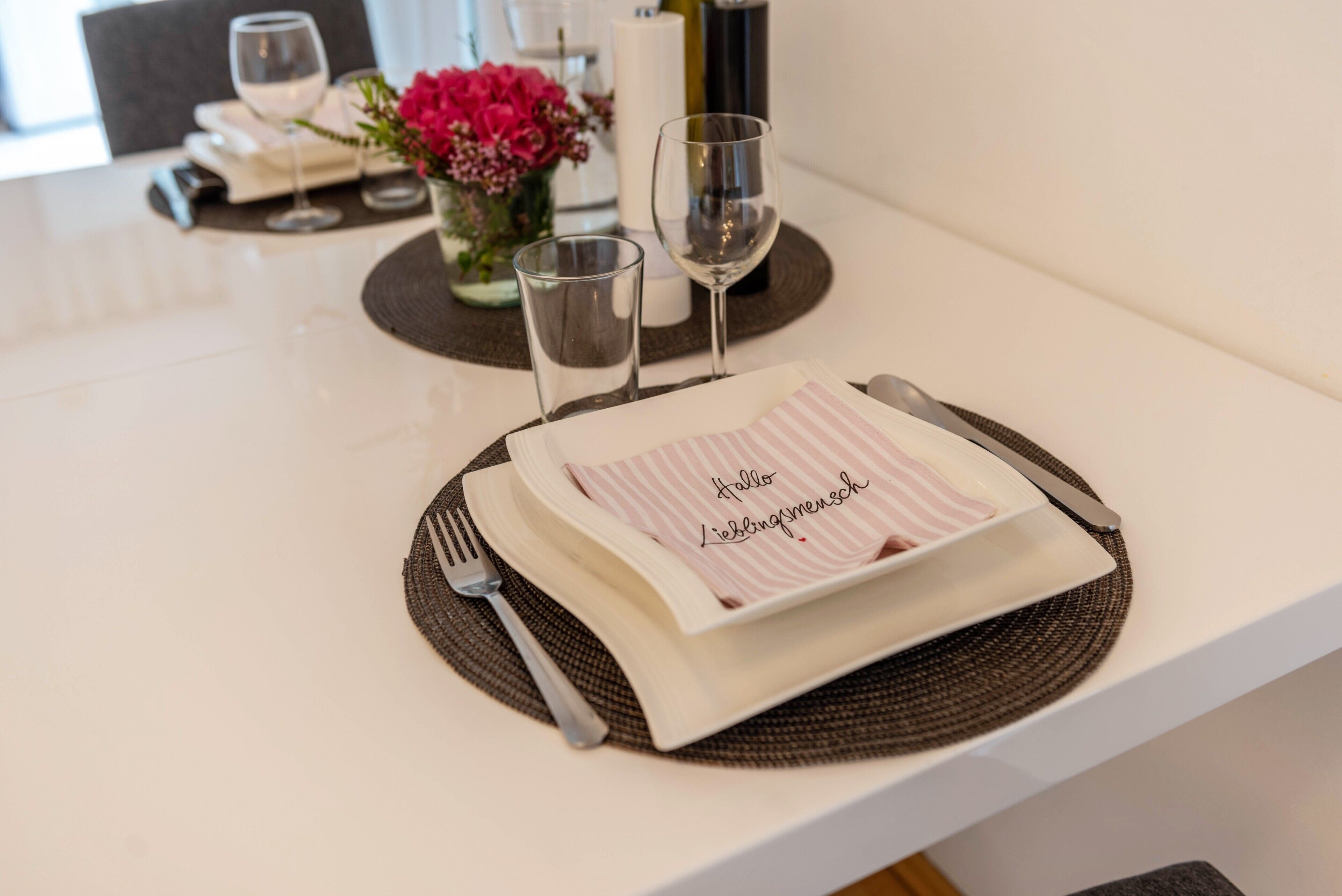 A table set with a placemat, a folded napkin with a message, a fork, a knife, and two wine glasses.