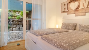 Une chambre avec un lit, une vue sur un jardin par une porte vitrée et un panneau indiquant « MAISON » sur le mur.