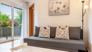 Un salon moderne avec un canapé gris, des coussins à motifs floraux, une table basse blanche ornée de bougies rouges et une œuvre d’art encadrée accrochée au mur.