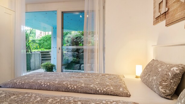 Une chambre avec un lit, une table de chevet, une fenêtre donnant sur un espace verdoyant et une porte menant à un espace extérieur.