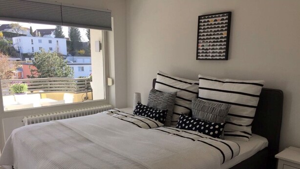 A bedroom with a bed, a nightstand, a window with a view of buildings, and a framed picture on the wall. - 'Saschas' Holiday Apartment with Lake View, Balcony, and Wi-Fi (Überlingen)
