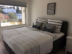A bedroom with a bed, a nightstand, a window with a view of buildings, and a framed picture on the wall. - Apartment 'Saschas' with Lake View, Balcony and Wi-Fi (Überlingen)