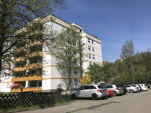A multi-story building with a white facade and yellow accents, a parking lot with several cars, and trees in the foreground. - Apartment 'Saschas' with Lake View, Balcony and Wi-Fi (Überlingen)