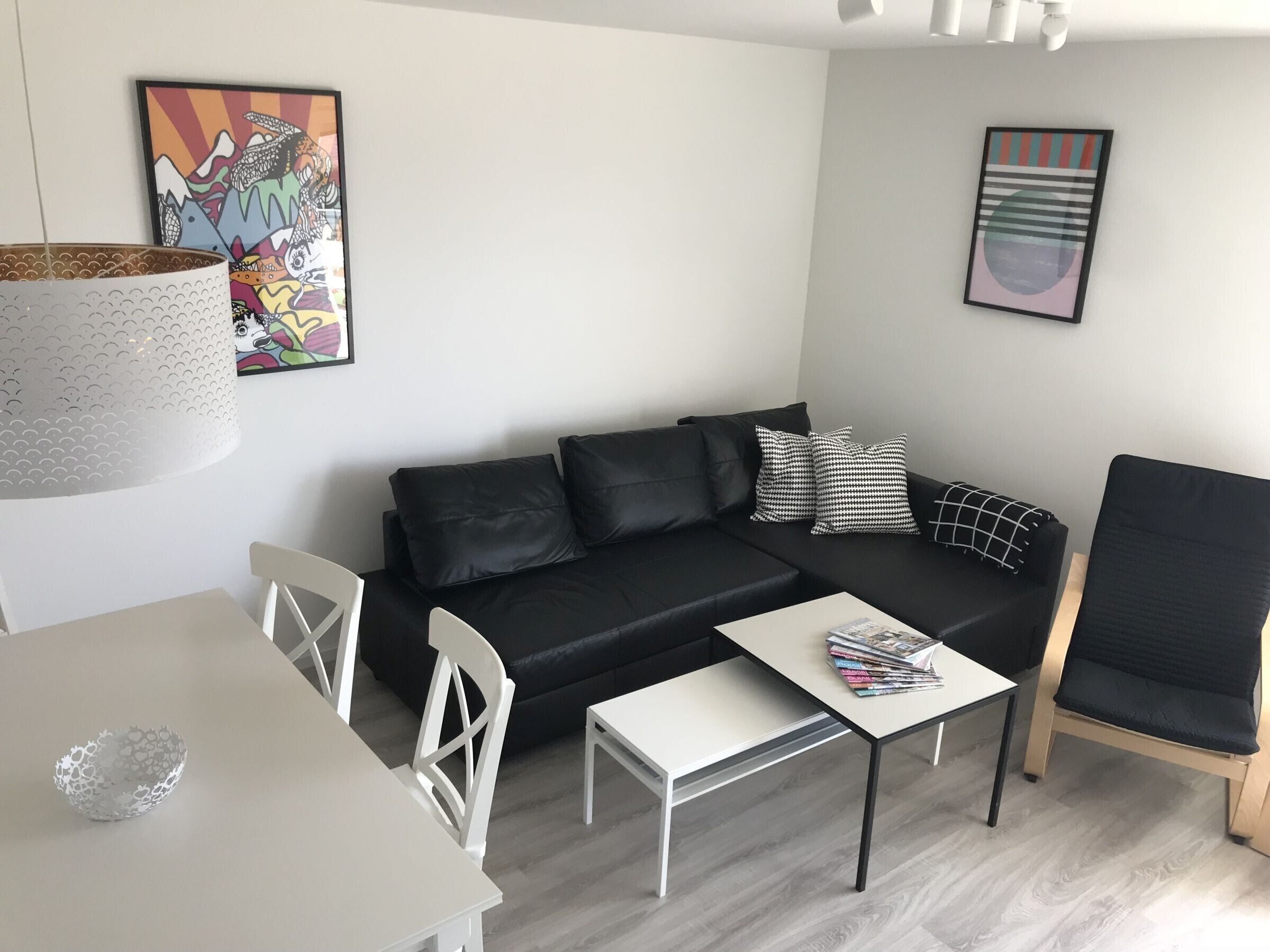 Un salon moderne avec un canapé en cuir noir, des chaises blanches, une petite table blanche et une table basse. Deux œuvres d’art sont accrochées au mur, et une lampe est suspendue au plafond.