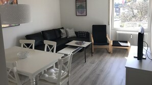 A modern living room with a black sofa, white dining table, and a black chair. There is a window with a view of the outside. - 'Saschas' Holiday Apartment with Lake View, Balcony, and Wi-Fi (Überlingen)