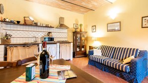 A cozy living room with a striped sofa, a dining table with a bottle of wine, and a kitchen area with a tiled backsplash and a stove. - Cottage in Tuscany with Pool & BBQ (San Casciano in Val di Pesa)