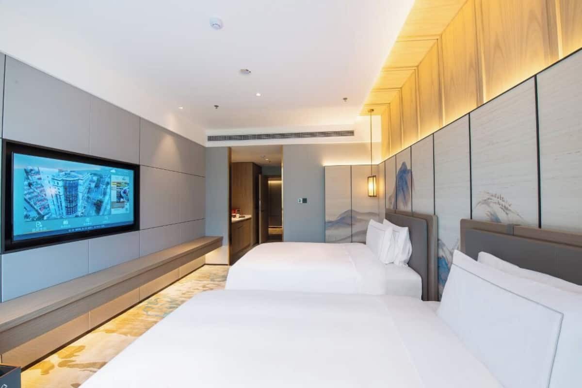 Ein Hotelzimmer mit großem Flachbildfernseher an der Wand, zwei Betten mit weißer Bettwäsche, einem Holz-Kopfschild und Blick auf eine gebirgige Landschaft.