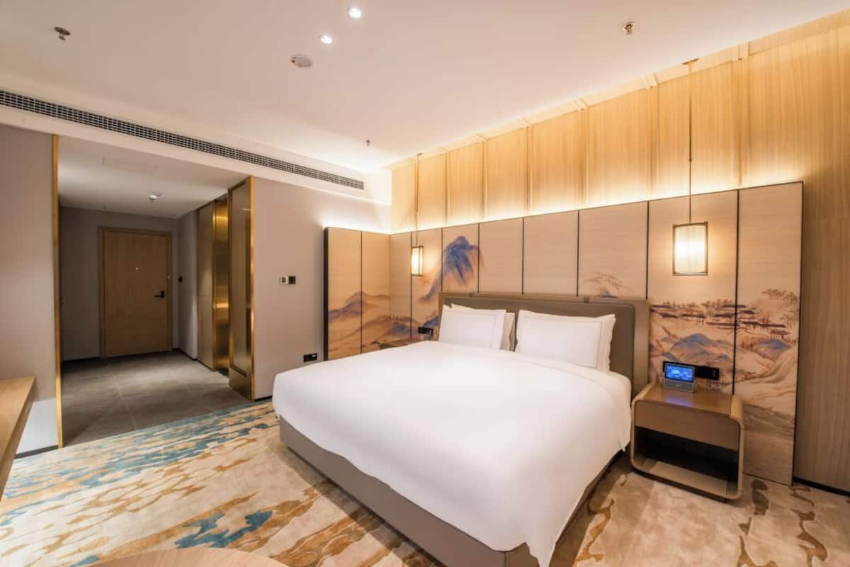 Ein Hotelzimmer mit einem großen Bett, einem Nachttisch mit digitaler Uhr und einem an der Wand befestigten Kunstwerk.