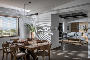 A modern living room with a dining area, featuring a large window, a potted plant, and a sofa with cushions.
