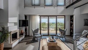 A spacious living room with a fireplace, two armchairs, a coffee table, and a large window offering a view of the outdoors. - Villa Ete: Prime 4BR Retreat with Private Pool (Stalida)