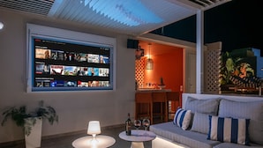 A modern living room with a large TV displaying Netflix content, a sofa with striped cushions, a small table with a lamp, and a dining area in the background.