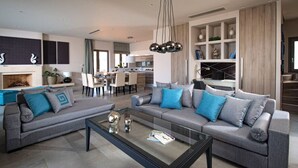 A modern living room with a grey sofa set, a glass coffee table, and a dining area with a white table and chairs. - Villa Belamar – Luxury Sea-View Stay, Hersonissos (Hersonissos)