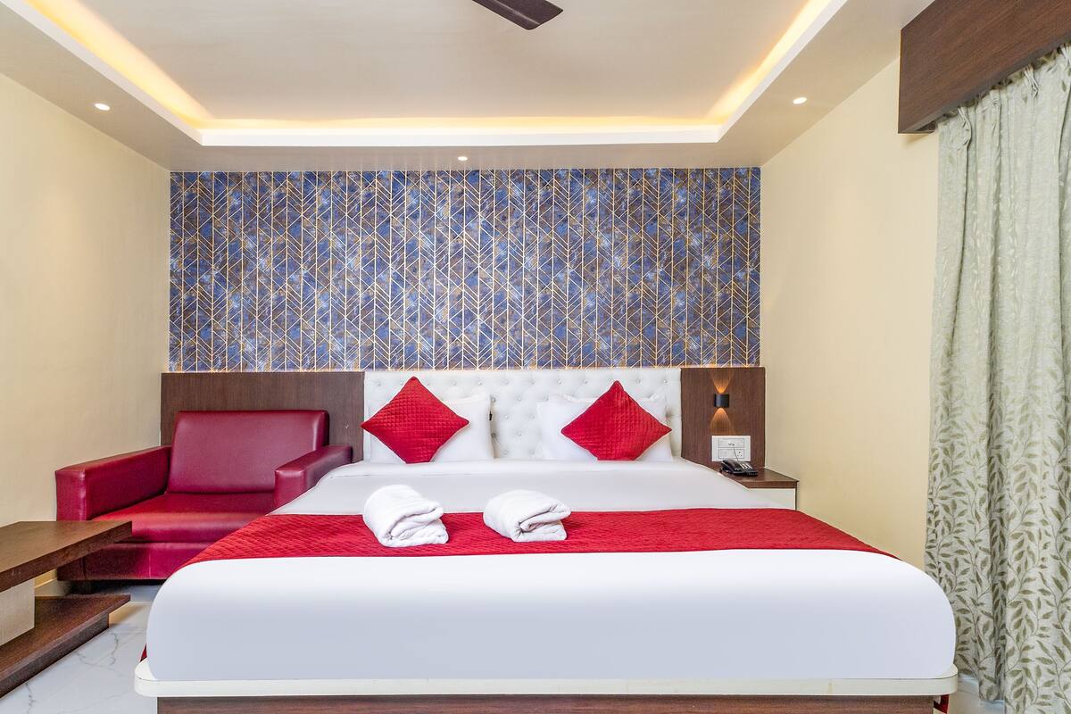 A hotel room with a large bed, red and white pillows, a red armchair, a wooden nightstand with a lamp, and a patterned wall.