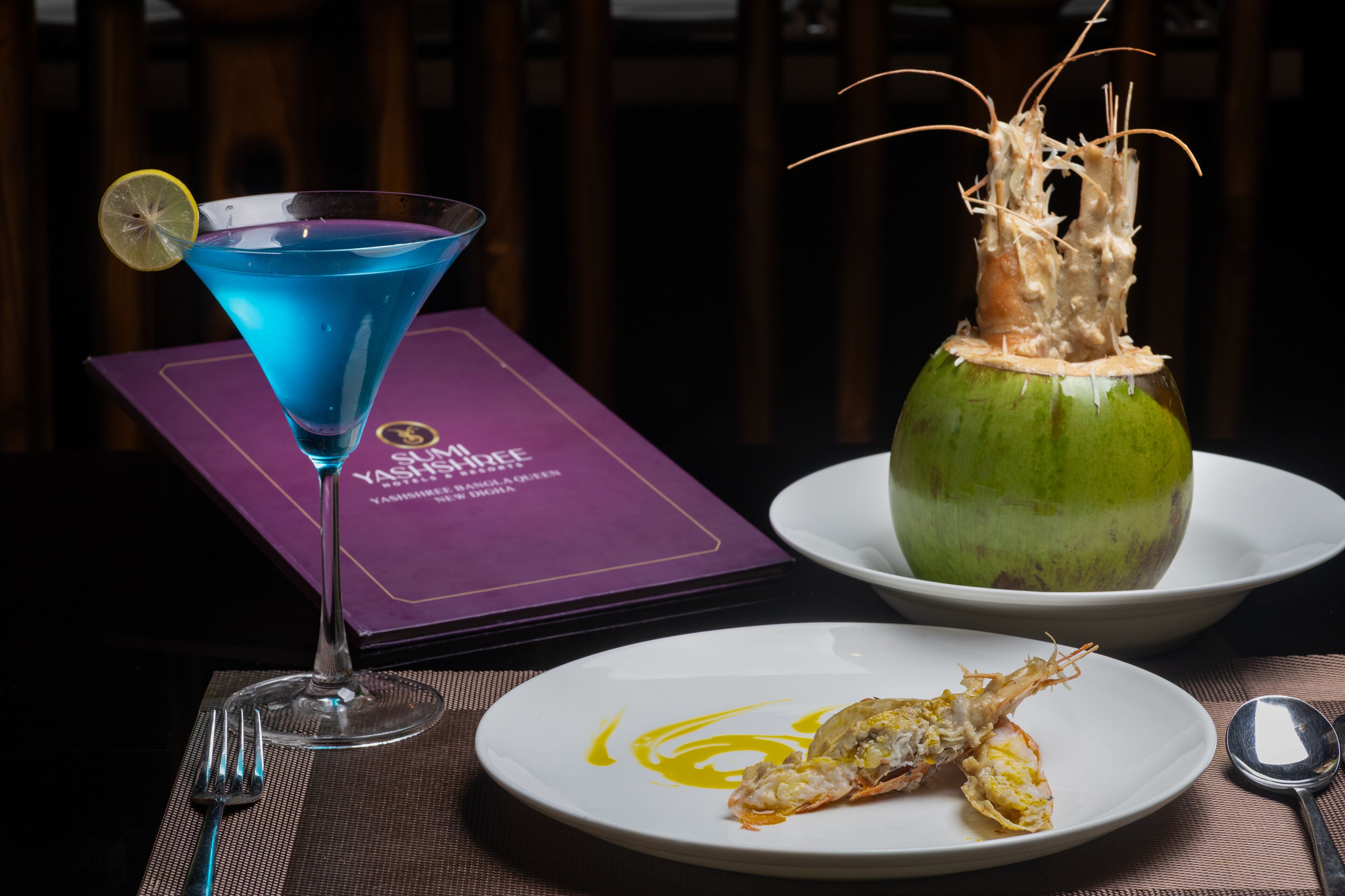 Een tafel met een blauwe cocktail, een bord met zeevruchten en een menukaart.