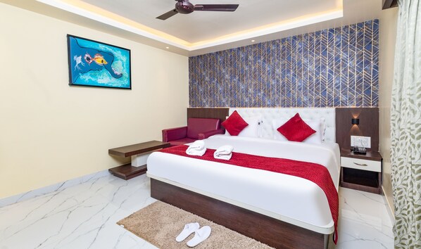 A modern hotel room with a large bed, red and white bedding, a red chair, a nightstand, and a ceiling fan. - Yashshree Bangla Queen (Kanthi)