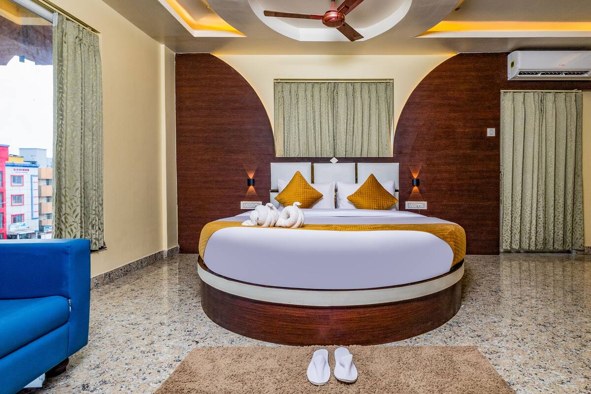 A hotel room with a round bed, a ceiling fan, an air conditioning unit, and a view of buildings outside.