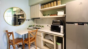 A compact kitchen with a round mirror, wooden chairs, a microwave, a toaster, and a refrigerator. - Poshtel Haven (Chiang Mai)