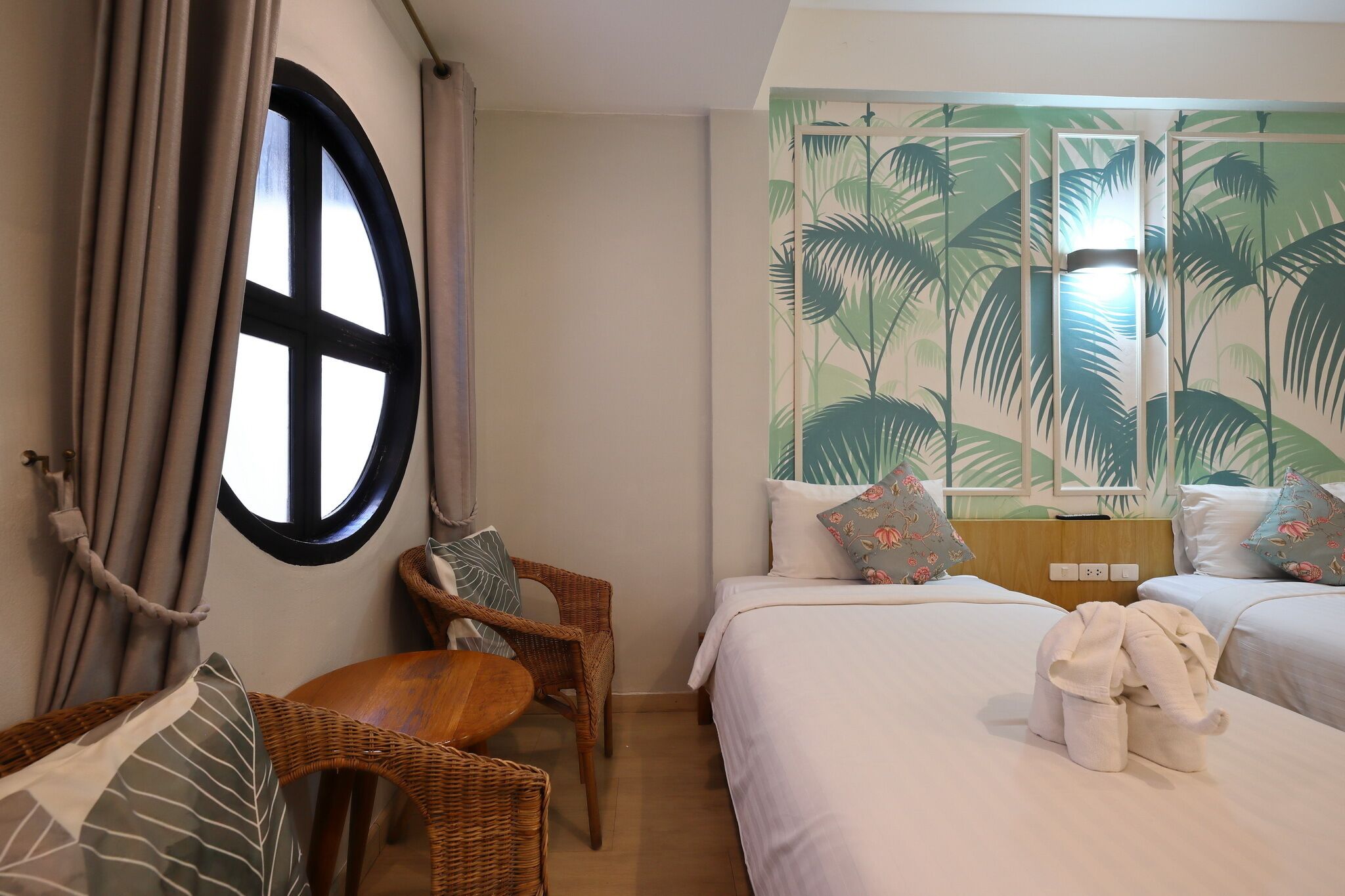 A hotel room with two beds, a round window, curtains, a wicker chair, and a wall with a tropical leaf pattern.