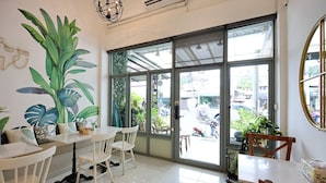 A cafe interior with white furniture, large windows, and a mural of tropical leaves. - Poshtel Haven (Chiang Mai)