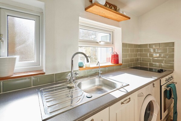 A kitchen with a stainless steel sink, a window, a washing machine, and a shelf with dishes. - Host Stay Baliol Street (Barnard Castle)