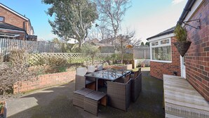 Een patio met een tafel met glazen blad en rieten stoelen, omgeven door een bakstenen muur en een houten terras.
