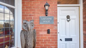 Een bakstenen muur met een bord waarop 'LOUVAINE HOUSE' staat, een witte deur met een beslagstuk en een houten uilbeeld.