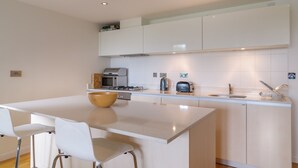 Una cocina moderna con armarios blancos, una isla central, estufa a gas, microondas, hervidor y tostadora. Hay dos sillas blancas en la isla.