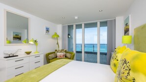Ein modernes Schlafzimmer mit einem großen Bett, einem grünen Sofa, einer weißen Kommode und einem Balkon mit Meerblick.