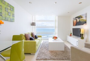 A modern living room with a view of the ocean, a green sofa, a white coffee table, and a flat-screen TV.