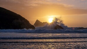 Un coucher de soleil sur un paysage côtier, avec des vagues qui s’écrasent contre des affleurements rocheux.