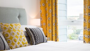 Un lit bien fait, agrémenté d’oreillers à motifs, d’une tête de lit rembourrée de couleur grise et de rideaux jaunes ornés de motifs floraux.