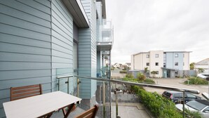 Un balcon avec une table et des chaises, une balustrade en verre, et une vue sur un parking et des bâtiments en arrière-plan.