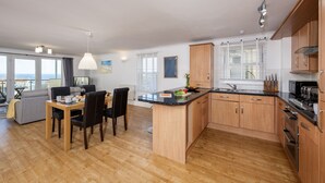 A modern kitchen with wooden cabinets, a dining area with a table and chairs, and a view of the city through large windows. - 8 Spinnakers, a charming two-bedroom apartment located in Pentire (Newquay)