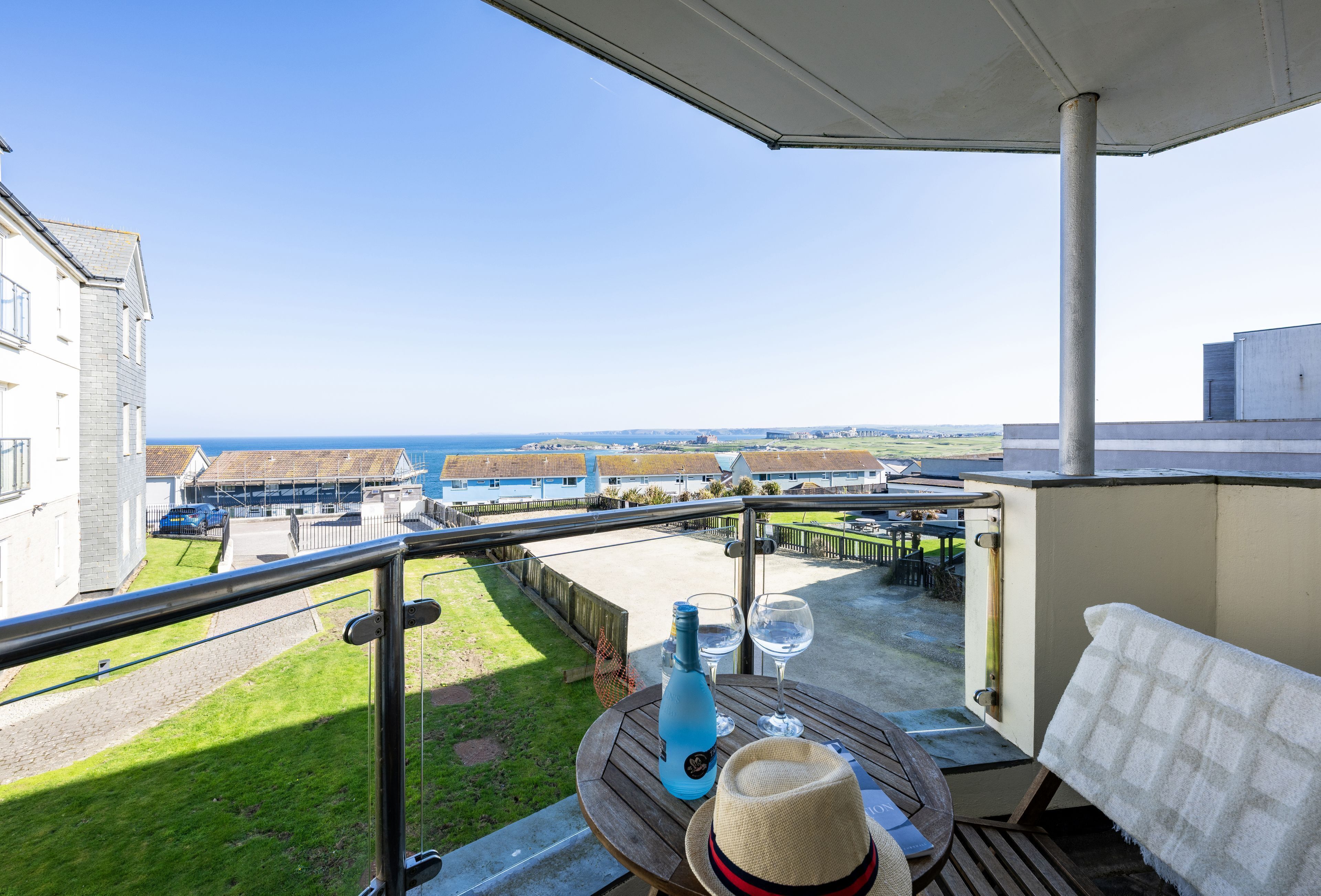 A balcony with a view of the sea, a bottle, two glasses, and a hat on a wooden table.