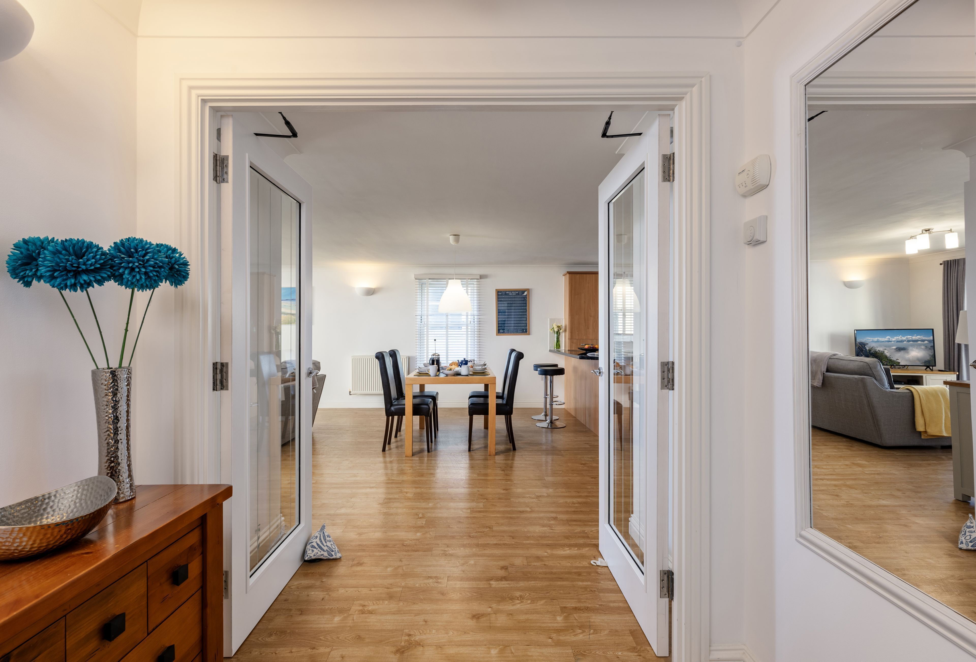 A modern interior with a dining area, wooden flooring, and a view into another room with a sofa and TV.