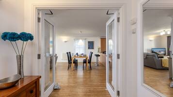 A modern interior with a dining area, wooden flooring, and a view into another room with a sofa and TV.