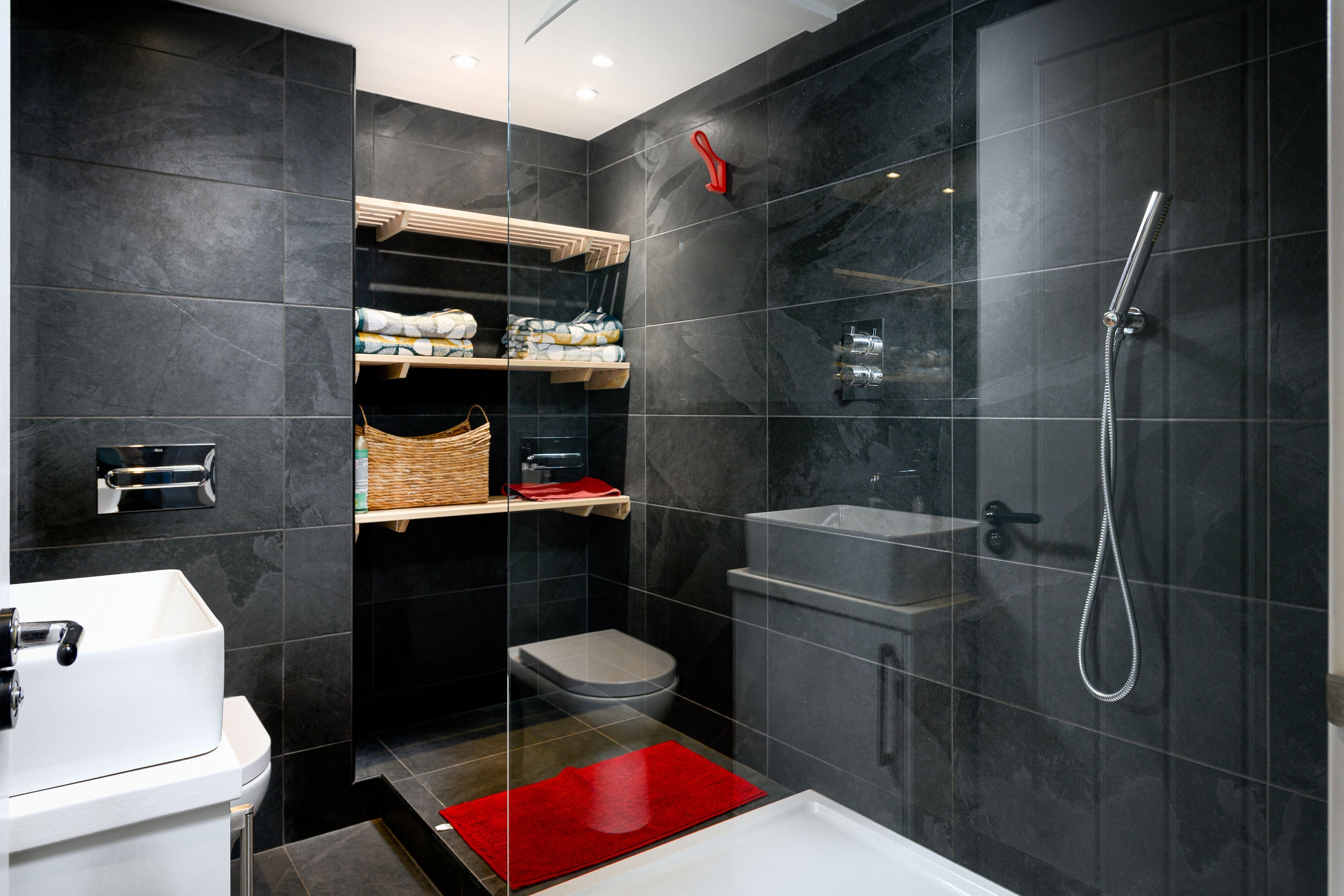 Une salle de bain moderne avec un lavabo blanc, des toilettes, une douche équipée d’un pommeau de douche à main et un tapis de bain rouge.