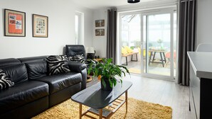 Una sala de estar moderna con un sofá de cuero negro, una mesa de centro y una planta en maceta. Hay cuadros enmarcados en la pared, una puerta corrediza de vidrio que da a un balcón y se puede ver un comedor al otro lado de la puerta.