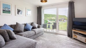 Ein modernes Wohnzimmer mit einem grauen Sofa, einem Flachbildfernseher und einem großen Fenster mit Blick auf eine grüne Landschaft.