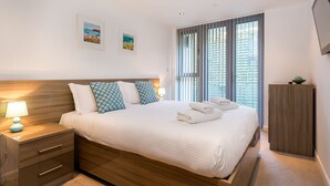 Una cama bien hecha con cabecera de madera, una mesita de noche con lámpara y dos cuadros en la pared.