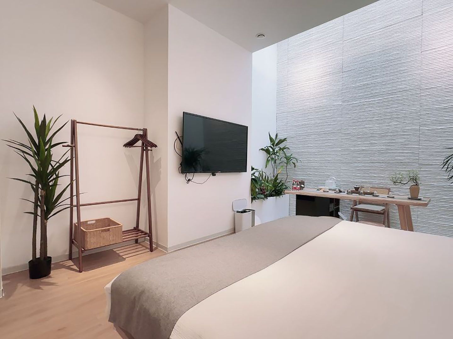 Ein modernes Schlafzimmer mit Bett, einem an der Wand montierten Fernseher, einem kleinen Schreibtisch mit Stuhl und einem Fenster mit Jalousien.