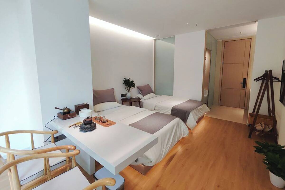 Ett modernt hotellrum med två sängar, ett skrivbord i trä, en stol, ett litet bord och en krukväxt.