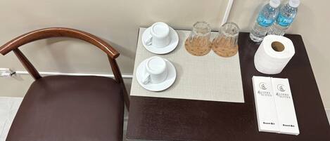 A hotel room with a brown leather chair, a small table with a placemat, two white cups, two glass tumblers, a roll of toilet paper, and two water bottles.