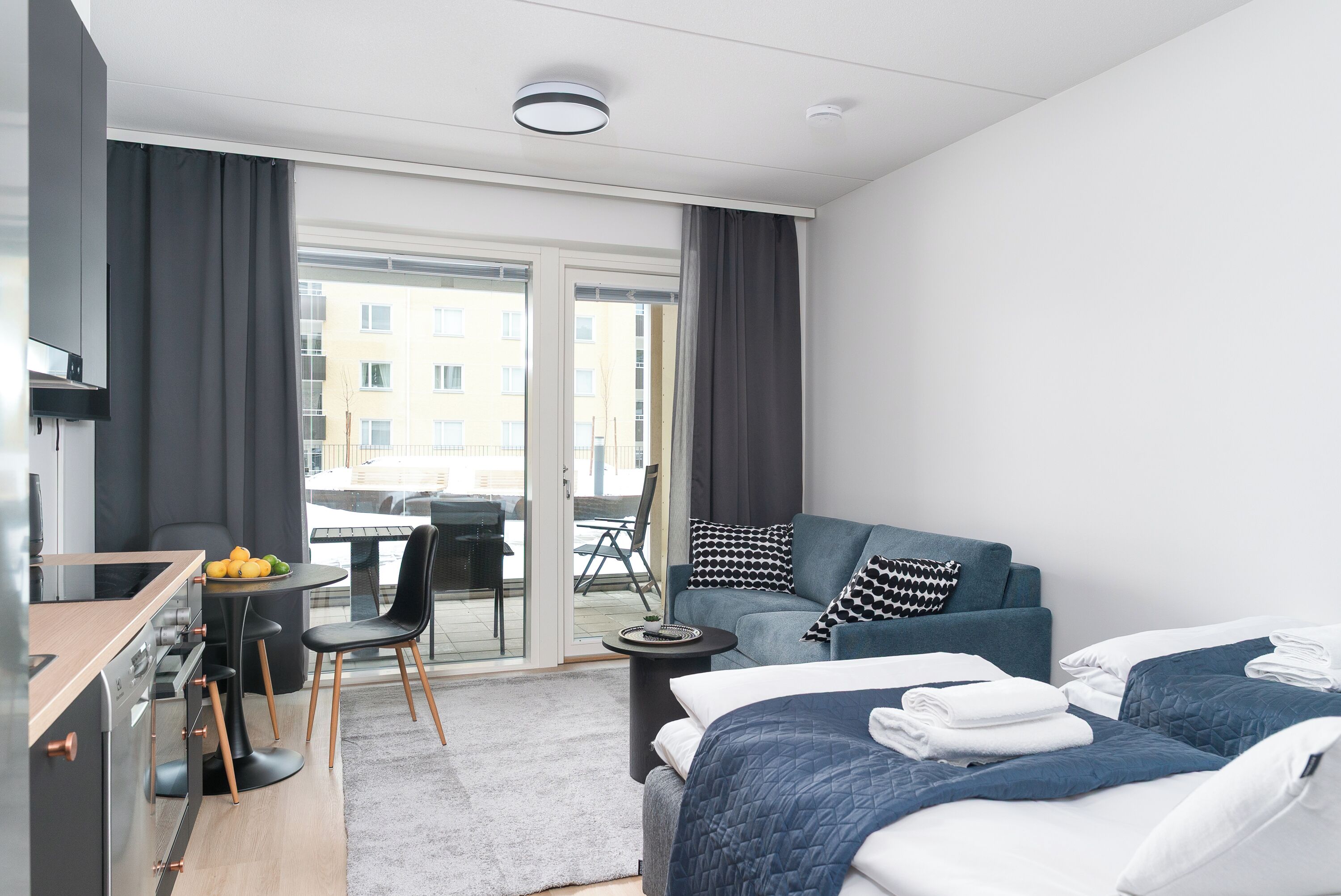 A modern hotel room with a kitchenette, a sofa bed, a dining area, and a view of the city.
