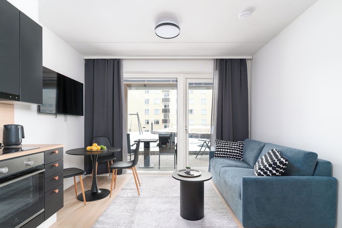 Un apartamento moderno con cocina, comedor y sala de estar. Cuenta con un televisor de pantalla plana, una mesa de comedor con sillas, un sofá con cojines estampados y una pequeña mesa redonda con un adorno.