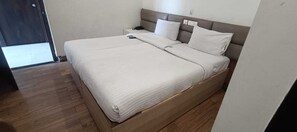 Una cama bien tendida con estructura de madera y ropa blanca, una mesita de noche con un teléfono y una puerta que da a un pasillo.