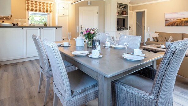 A dining area with a table set for four, surrounded by wicker chairs. A kitchen with white cabinets is visible in the background. - Bright 3-room apartment with approx. 70 m² for up to 4 people (Westerland)